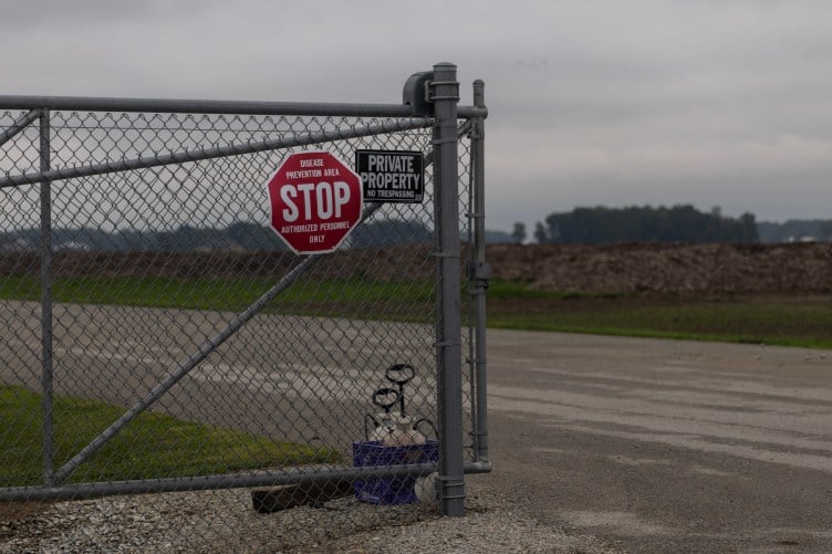 A stop sign reading, “Disease prevention area. Stop. Authorized personnel only,” hangs on a chain-link gate along an empty road. Across the road are several large mulch-covered mounds in a field.