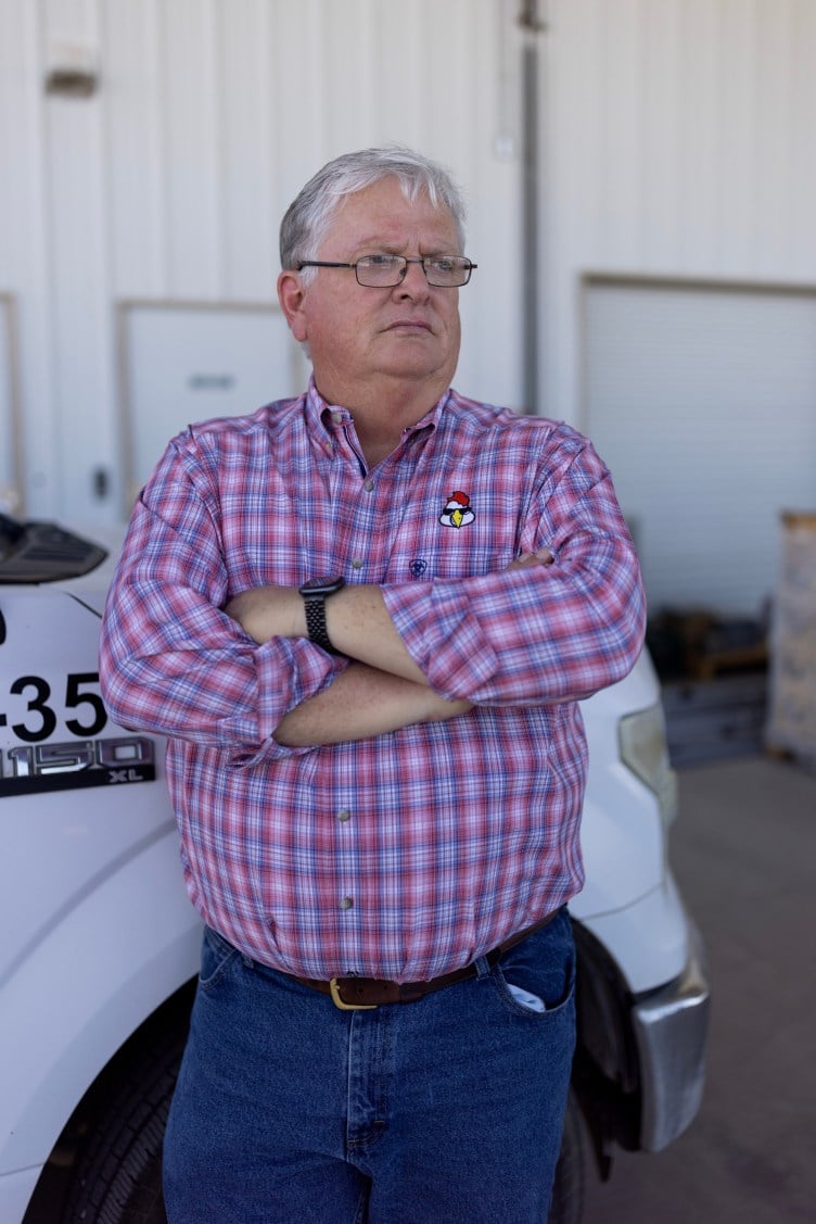 A man with white hair and glasses, wearing a plaid shirt with an embroidered chicken above the breast pocket, folds his arms in front of him, leaning against a white truck. 