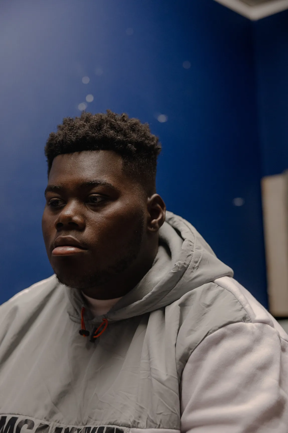 A teenage boy in a blue room looks solemnly off camera.