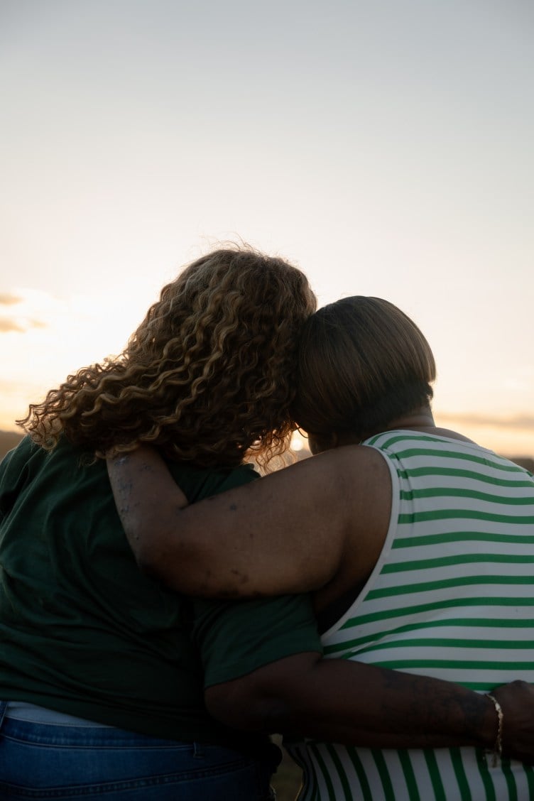 Two women, seen from behind, wrap their arms around each other and look toward the sunset.