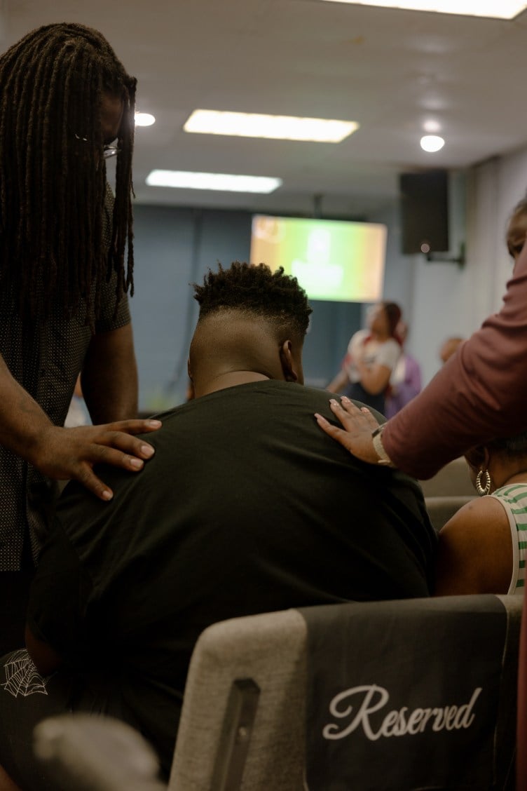 A teen boy is seated in a chair labeled “reserved.” Two women stand on either side of him with their heads bowed and hands resting on his shoulders.