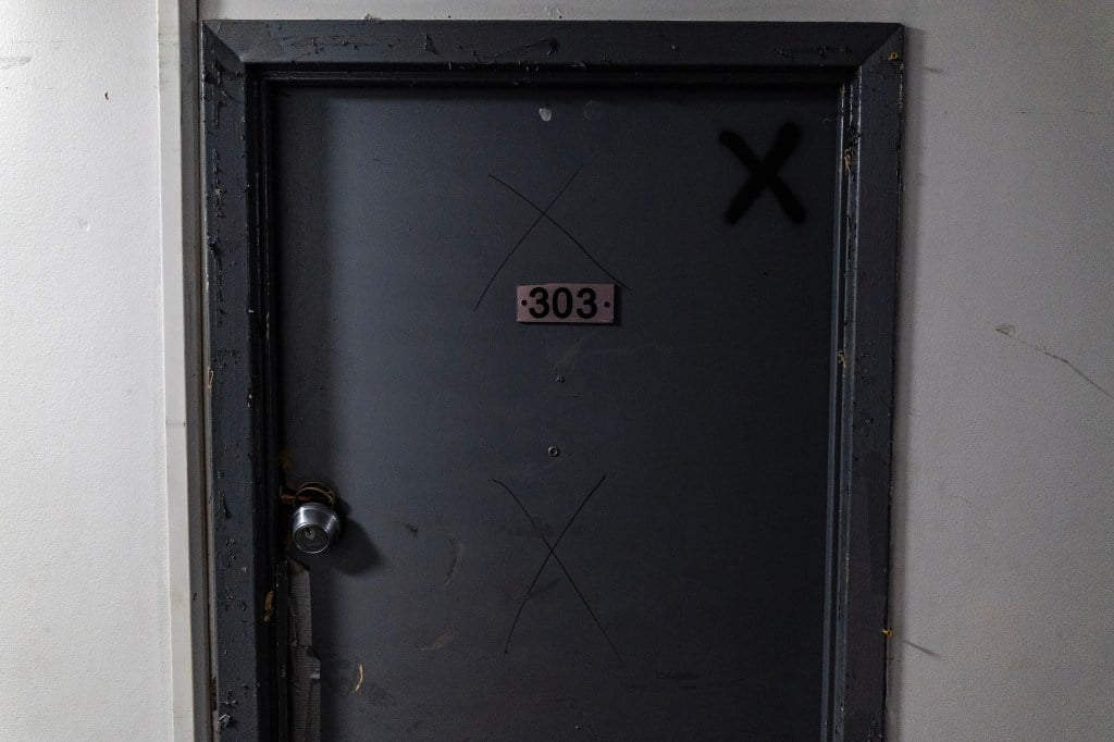 A door with three X’s drawn on it. The doorknob is broken and dangling.