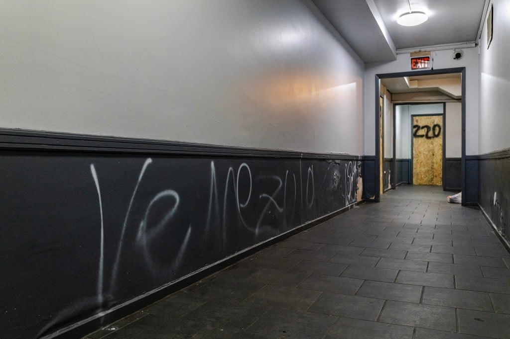 A hallway with “Venezula” spray painted in white on the left wall. At the end of the hallway is a door covered in plywood with “220” spray painted on it.