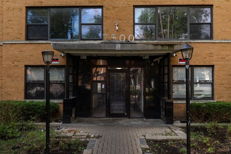 The front entrance of an apartment building with the numbers 7500 on an awning. The walkway is made of mismatched materials. The blinds seen through the right side windows are broken. On either side of the entrance door there are “No Trespassing" signs.