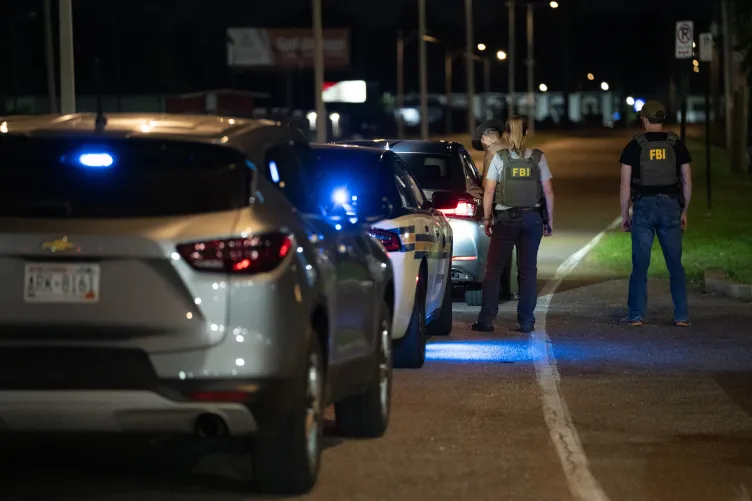 A person with a wide-brimmed hat leans into a stopped car. Two other people stand near him, wearing vests with “FBI” written on them.