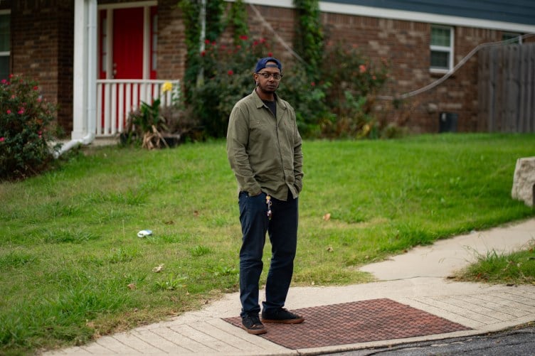 A Black man stands on the sidewalk in front of a house and lawn.