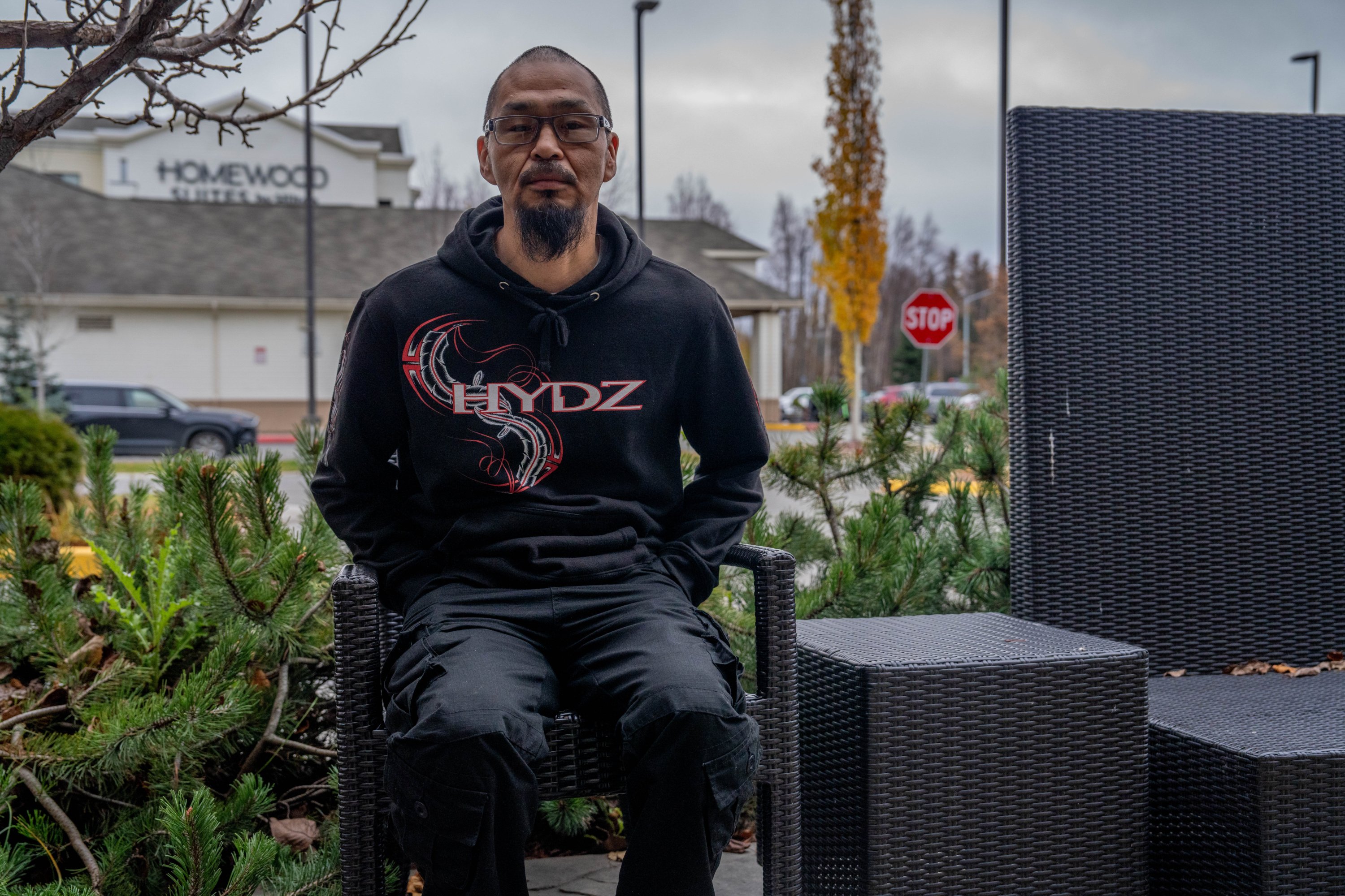 Draußen sitzt ein Mann mit Spitzbart und Brille auf einem Korbstuhl. Hinter ihm sind ein Homewood Suites-Schild und ein Parkplatz zu sehen.