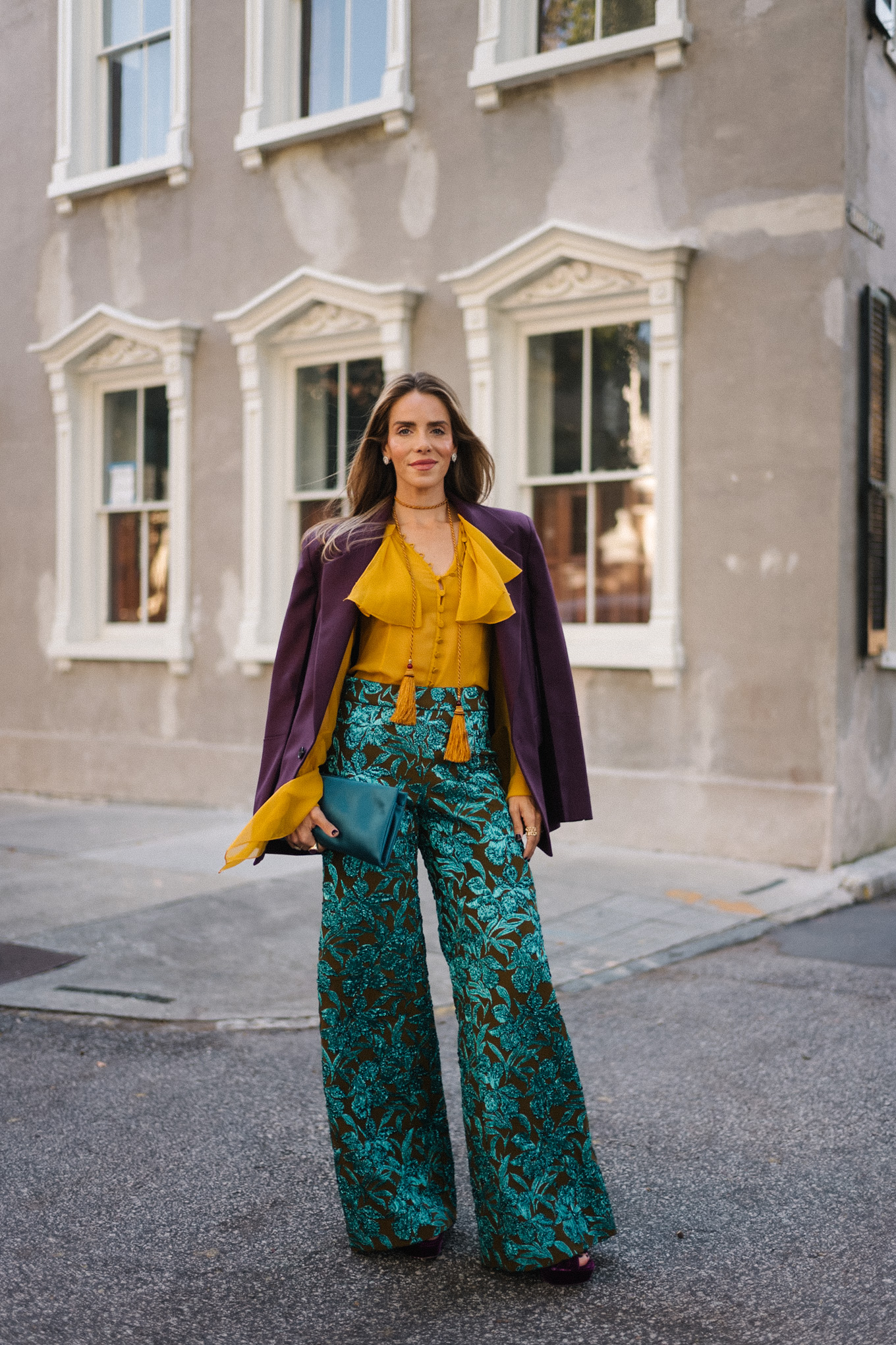 Blazer prune, chemisier à volants dorés, pantalon jacquard, pochette turquoise, escarpins violets