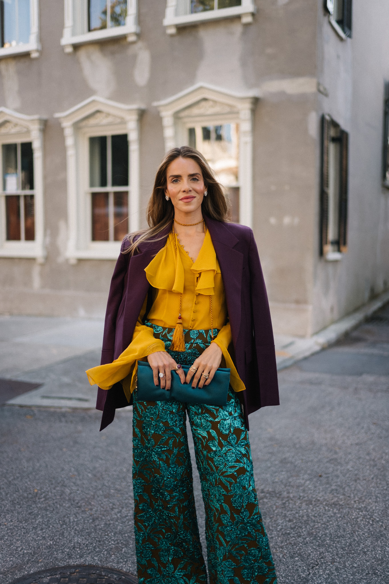 Blazer prune, chemisier à volants dorés, pantalon jacquard, pochette turquoise, escarpins violets