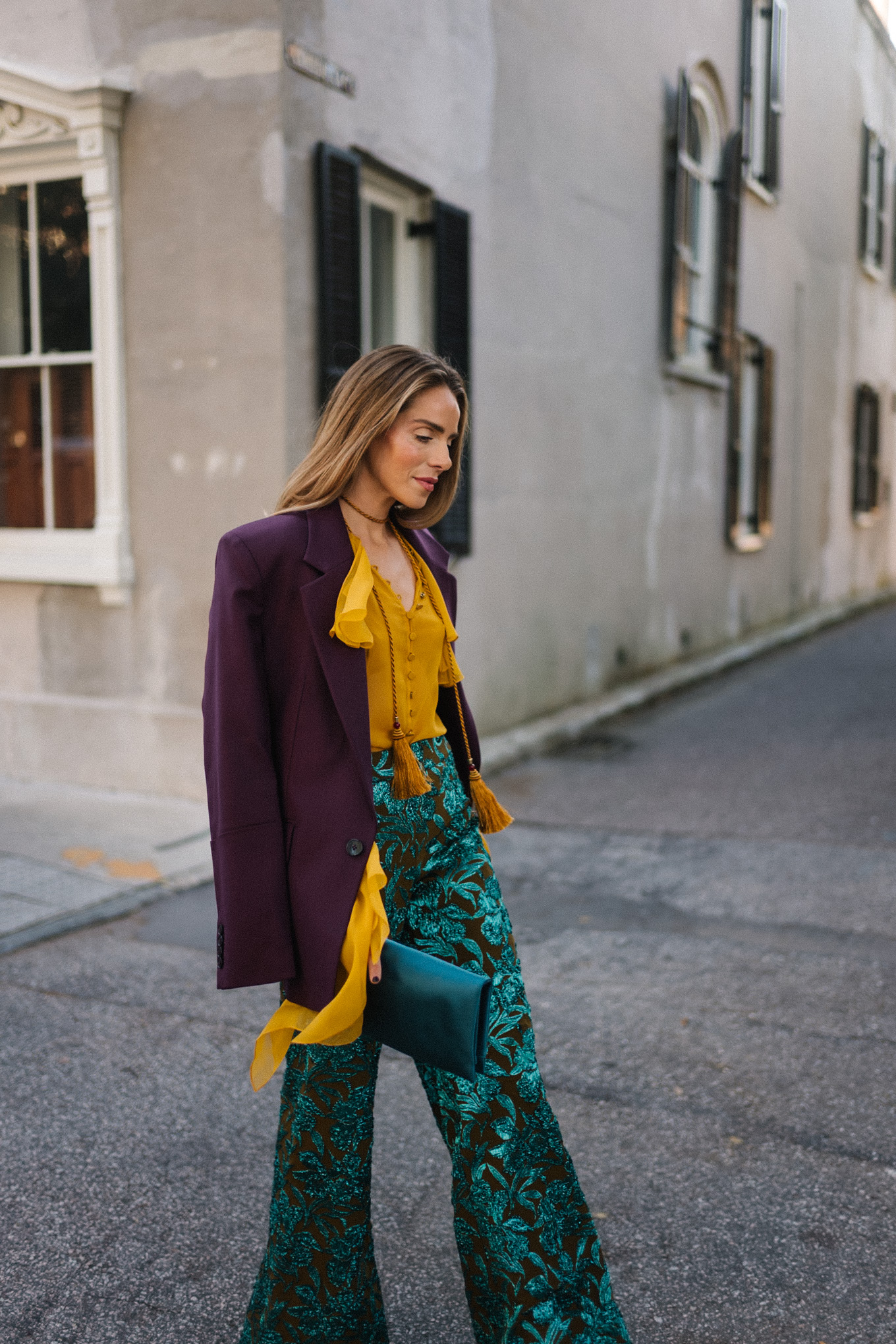 Blazer prune, chemisier à volants dorés, pantalon jacquard, pochette turquoise, escarpins violets