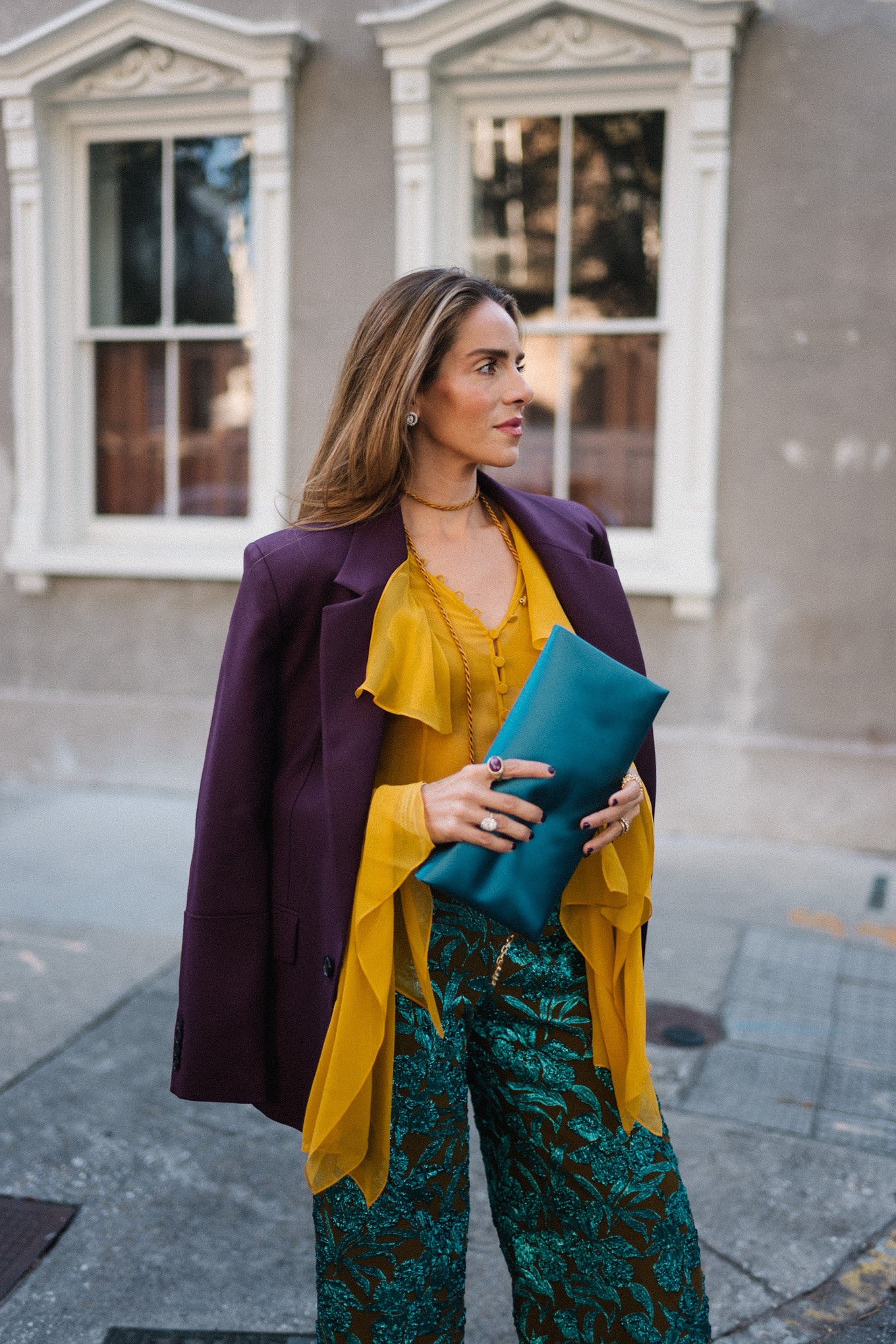 Blazer prune, chemisier à volants dorés, pantalon jacquard, pochette turquoise, escarpins violets