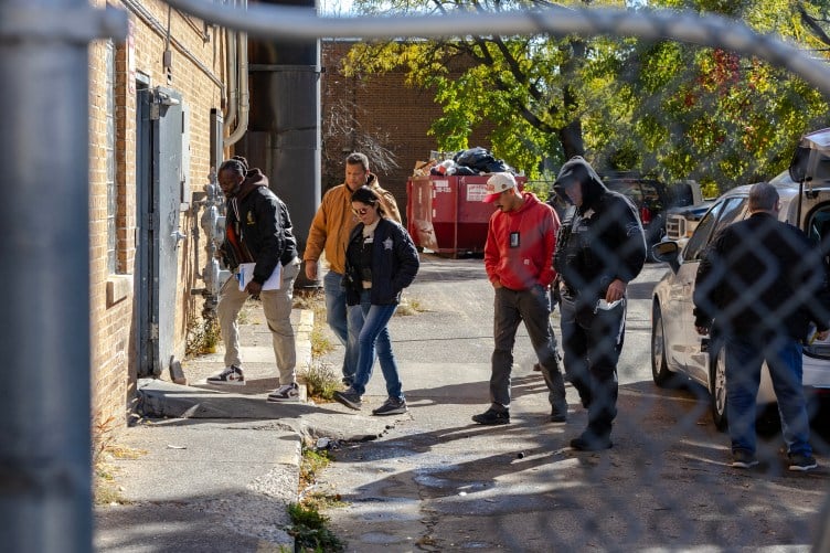 A group of people walking into the back door of a building. One person is carrying a clipboard and two of them are wearing badges.
