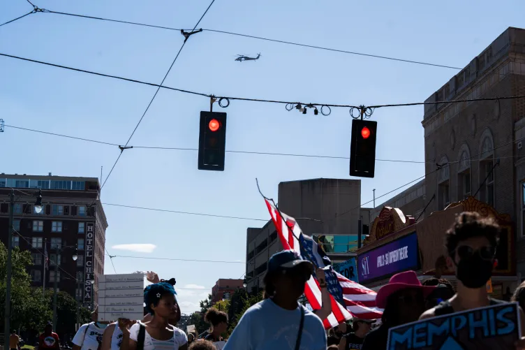 A crowd of people hold protest signs and an American flag. A helicopter is in the distance above them.