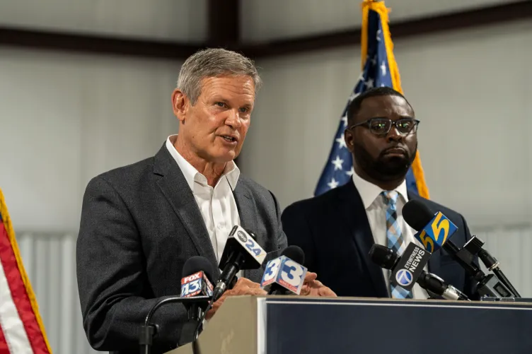Two men wearing suits stand at a podium. One of them speaks into numerous news station microphones.