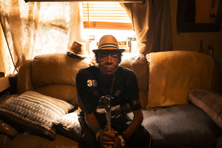 A man holding a cane and wearing a fedora sits on a sofa.