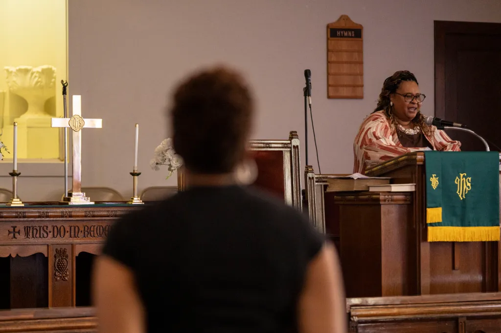 A woman stands at the front of a church speaking into a microphone near the altar and a cross. One out-of-focus person faces her in the foreground.