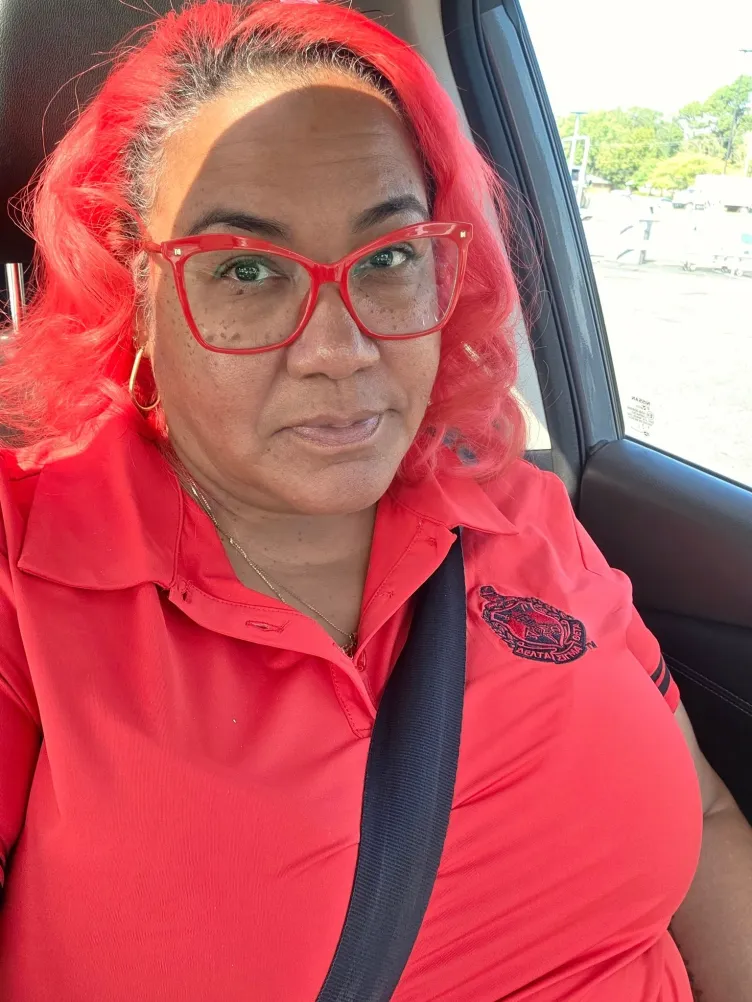A Black woman in a selfie-style photograph sits in a car with a seat belt across her torso.