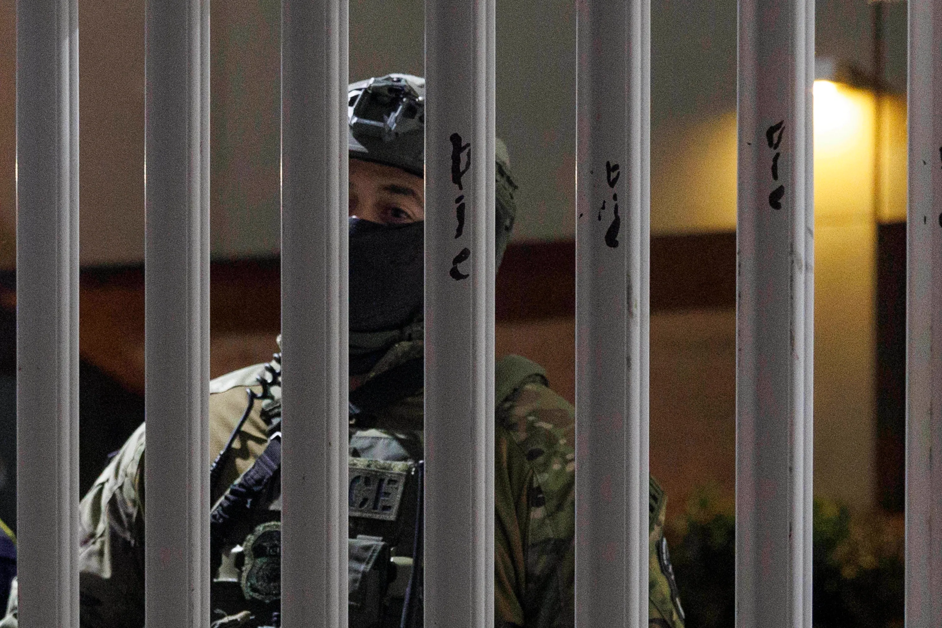A masked ICE agent wearing a helmet and camouflage uniform looks through bars that have the words “Die, die, die” written on them.