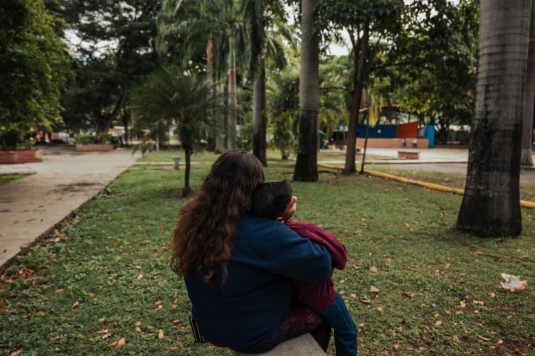 Une femme et un enfant portant des survêtements bleus et marron sont assis face à la caméra sur une plage dans une pelouse d'un parc urbain avec de grands palmiers et d'autres plantes tropicales luxuriantes.