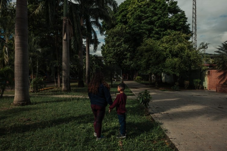 A woman and child wearing blue and maroon sweatsuits stand facing away from the camera in a grassy area of a city park, with tall palm trees and other lush tropical plants.