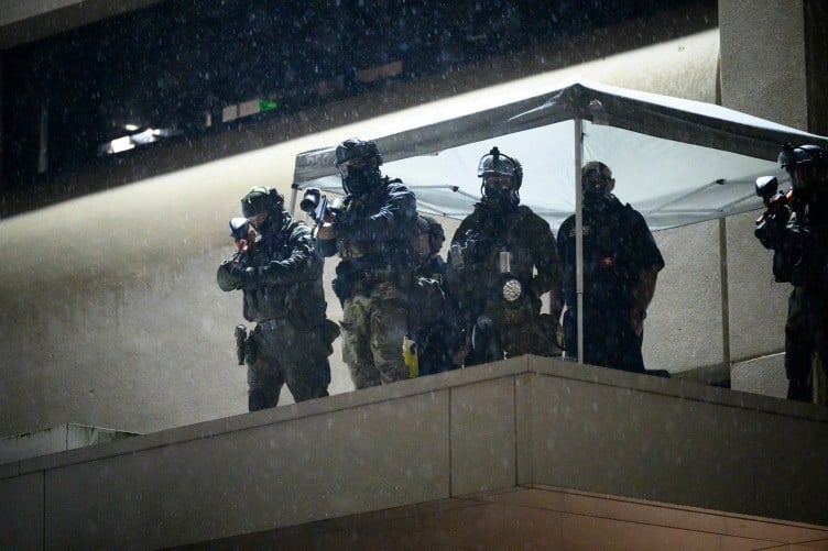 A group of around six agents wearing camouflage, masks and helmets stand on an elevated surface. It is raining and they are underneath a white tent. Three of them are pointing gun-like weapons toward the scene below them.