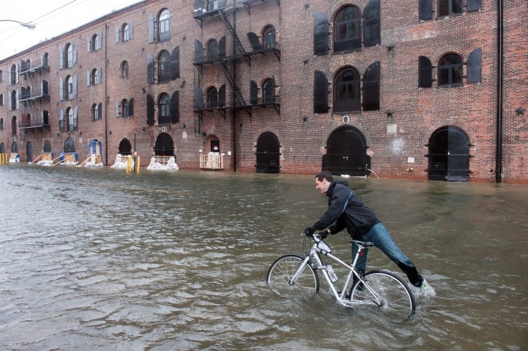 A man gets onto a bicycle while standing in water that is almost up to his knees in front of a brick building with black shutters.
