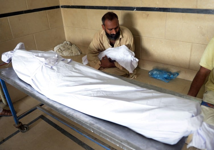 A man cries while tenderly cradling a small body wrapped in white fabric. In front of him is an adult-sized body also wrapped in white fabric.