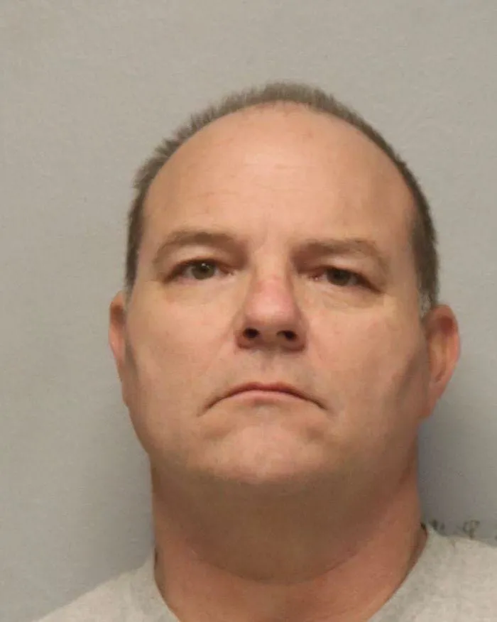 A booking photo of an older white man with balding hair and a cleanly shaven face, wearing a white T-shirt. 