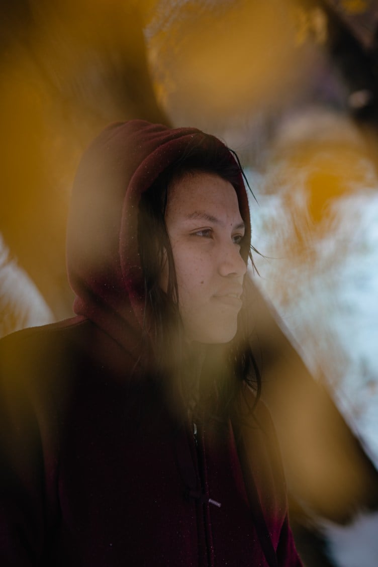 A woman with dark hair escaping from the hood of a maroon sweatshirt stares to the right. Golden leaves, out of focus in the foreground, dot the image and slightly blur parts of her face.
