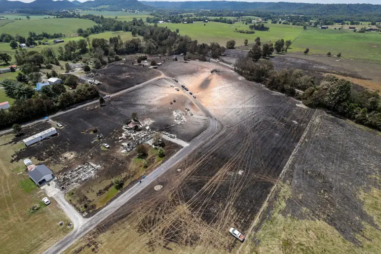 A large area of rural land seen from above. Most of the area is covered in green grass but a circular concentration of the land is stained black and within that area are burned buildings.