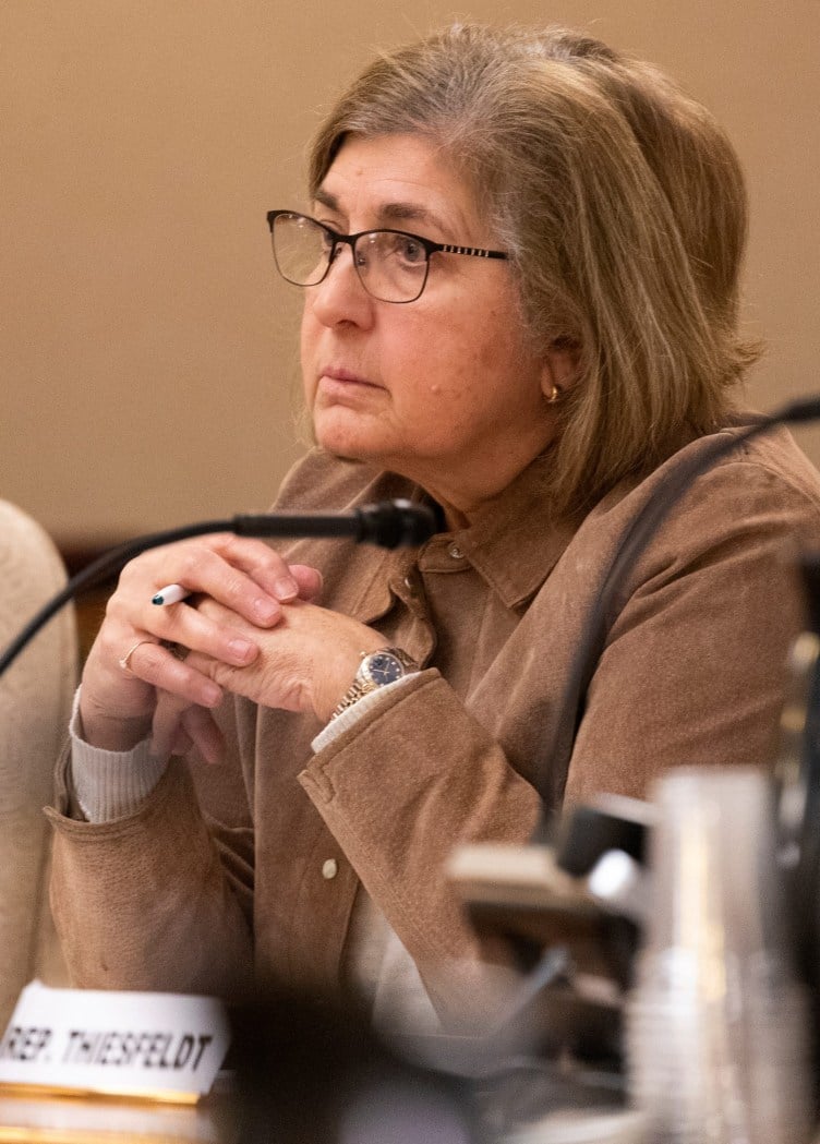 Eine Frau mit Brille und braunem Haar, gekleidet in eine braune Jacke, sitzt an einem Tisch.