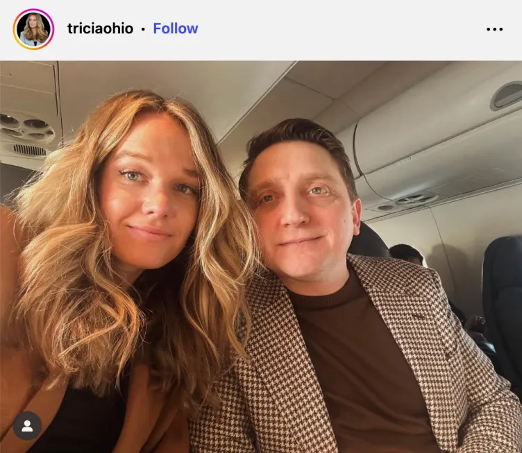 Eine Frau mit blonden Haaren posiert mit einem Mann, der eine braune Anzugjacke mit Hahnentrittmuster trägt, für ein Selfie im Flugzeug.