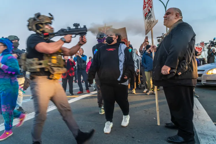 An agent wearing a helmet and a camouflage vest fires a weapon at Jorge Bautista, who is standing a few feet in front of him holding a sign. The weapon is emanating smoke. Behind them there are protesters wearing plain clothing.