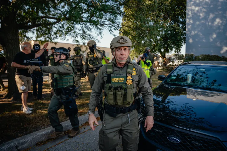 Greg Bovino wearing a helmet, vest and green uniform walking with agents behind him. One agent is pointing off to the left. There are people in plain clothes around the agents.