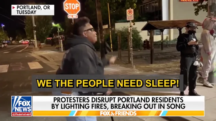 A person on a street with a cane shaking their hand at two people, one of whom is wearing a unicorn costume, above the words: “We the people need sleep!” A chyron shows the text: “Protesters disrupt Portland residents by lighting fires, breaking out in song.”