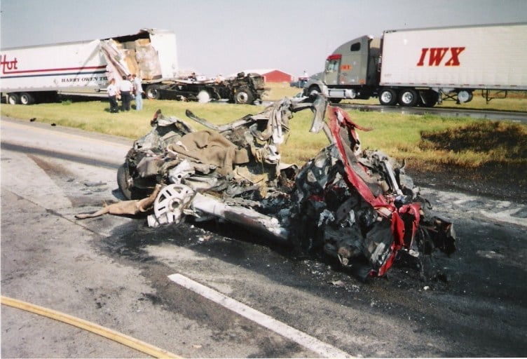 In the foreground there are the remnants of a car. Barely any elements of the car are recognizable and all that remains is a pile of metal. In the background there are two semitrucks. The truck on the left has some of its siding peeled off.