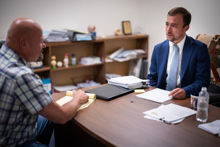 Un homme aux cheveux courts, vêtu d'un costume bleu et d'une cravate, discute avec un homme chauve dans un bureau, des documents étalés sur la table entre eux.