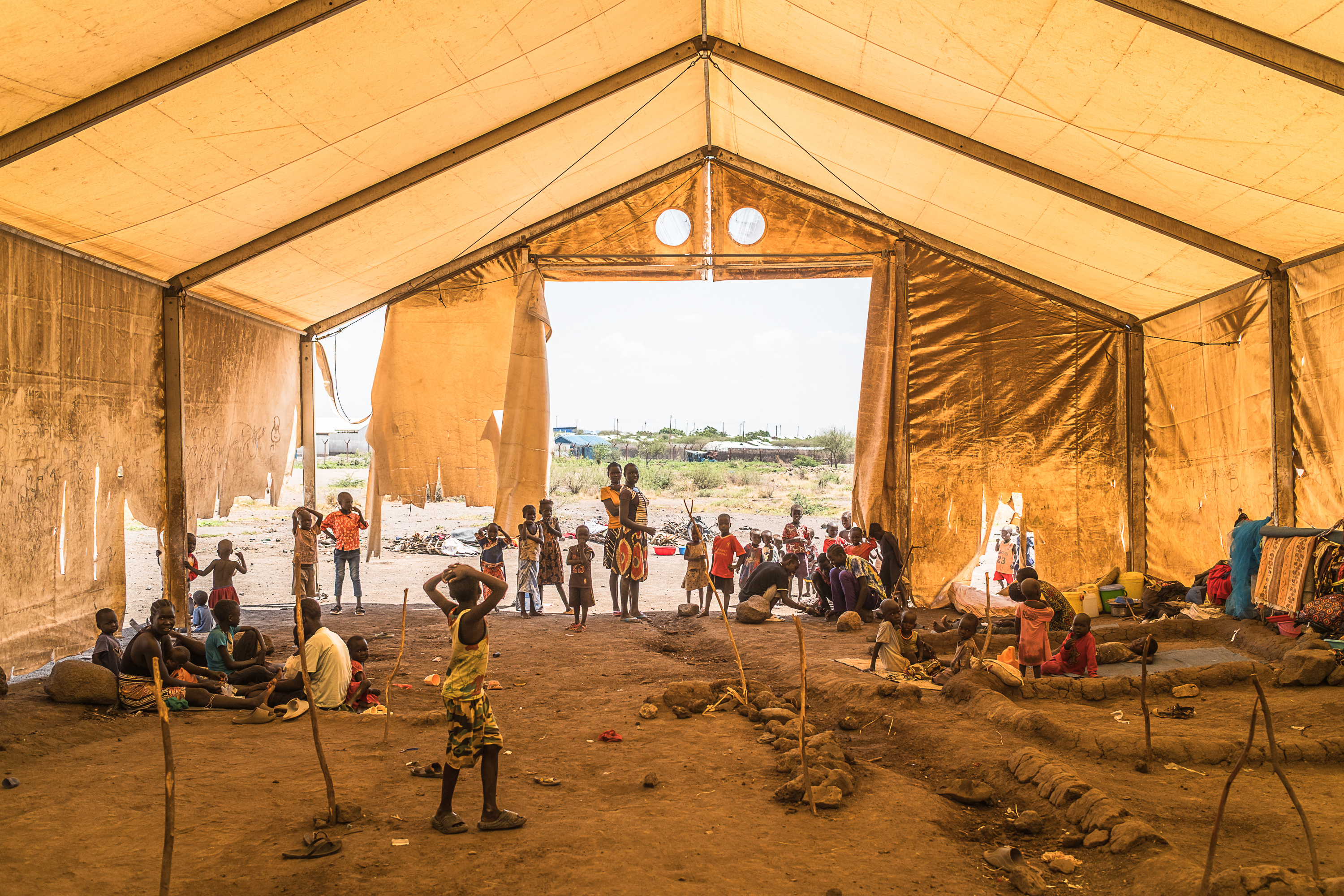 Under a large brown tent, people mill about, playing, conversing, and resting. Rocks and sticks on the ground mark sleeping areas for people and families.