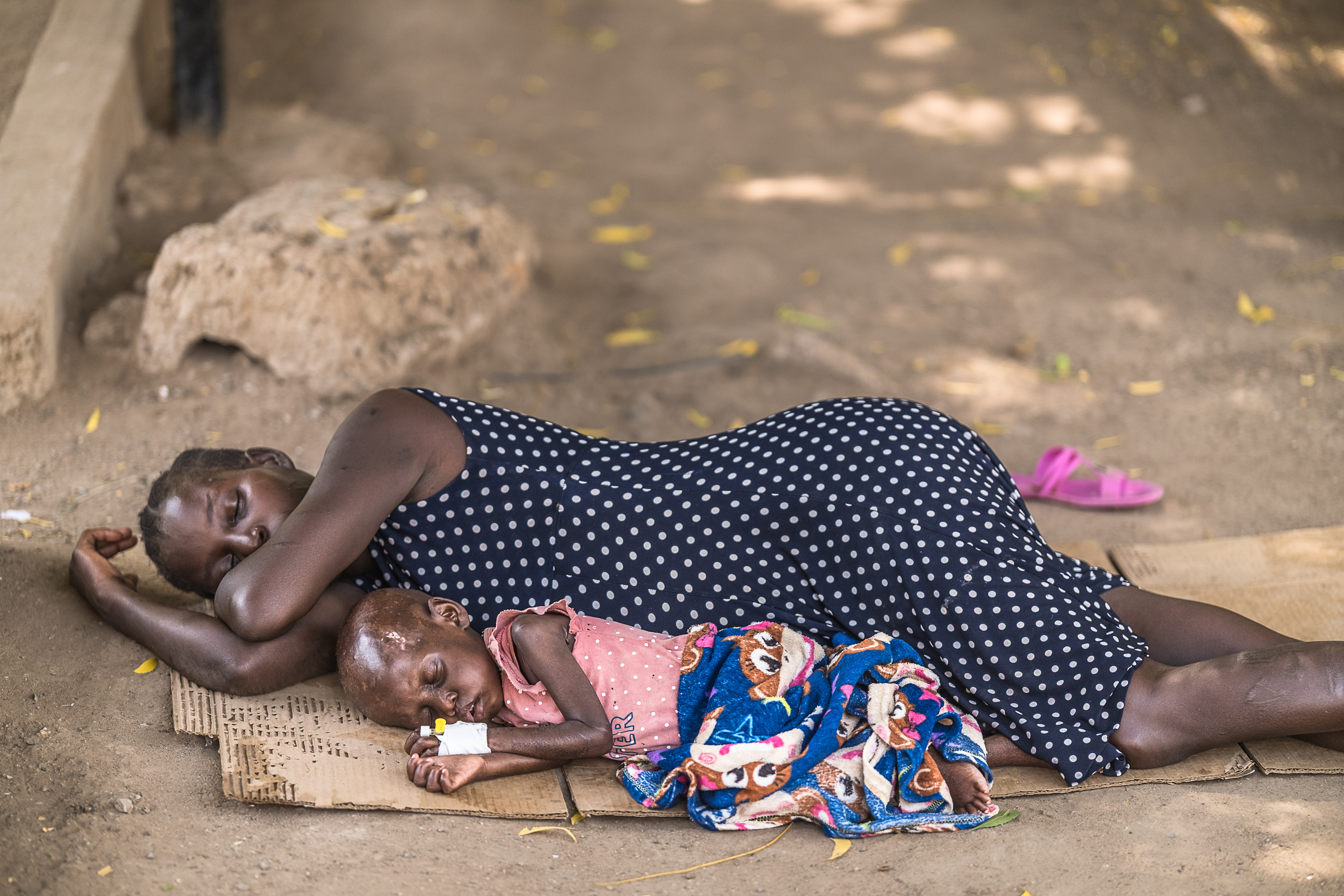 A woman in a polka-dot dress lies on her side on a piece of cardboard on the dusty earth. Her child, emaciated and with an IV port still bandaged on his left hand, nestles against her. A blanket with cartoon characters covers his thin legs.