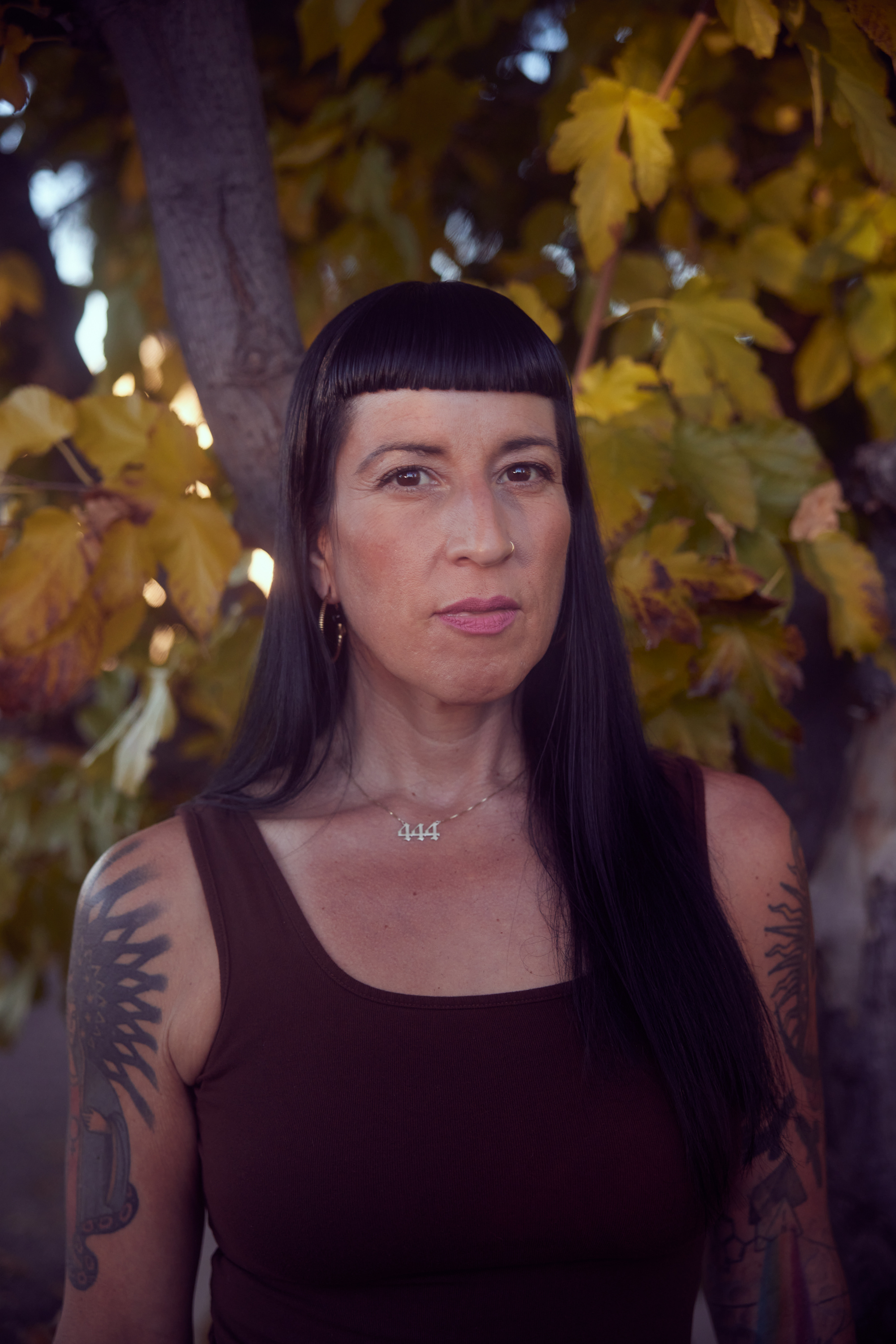 A woman with long dark hair, short bangs and tattoos, wearing a tank top, stands for a portrait in front of a mesquite tree.