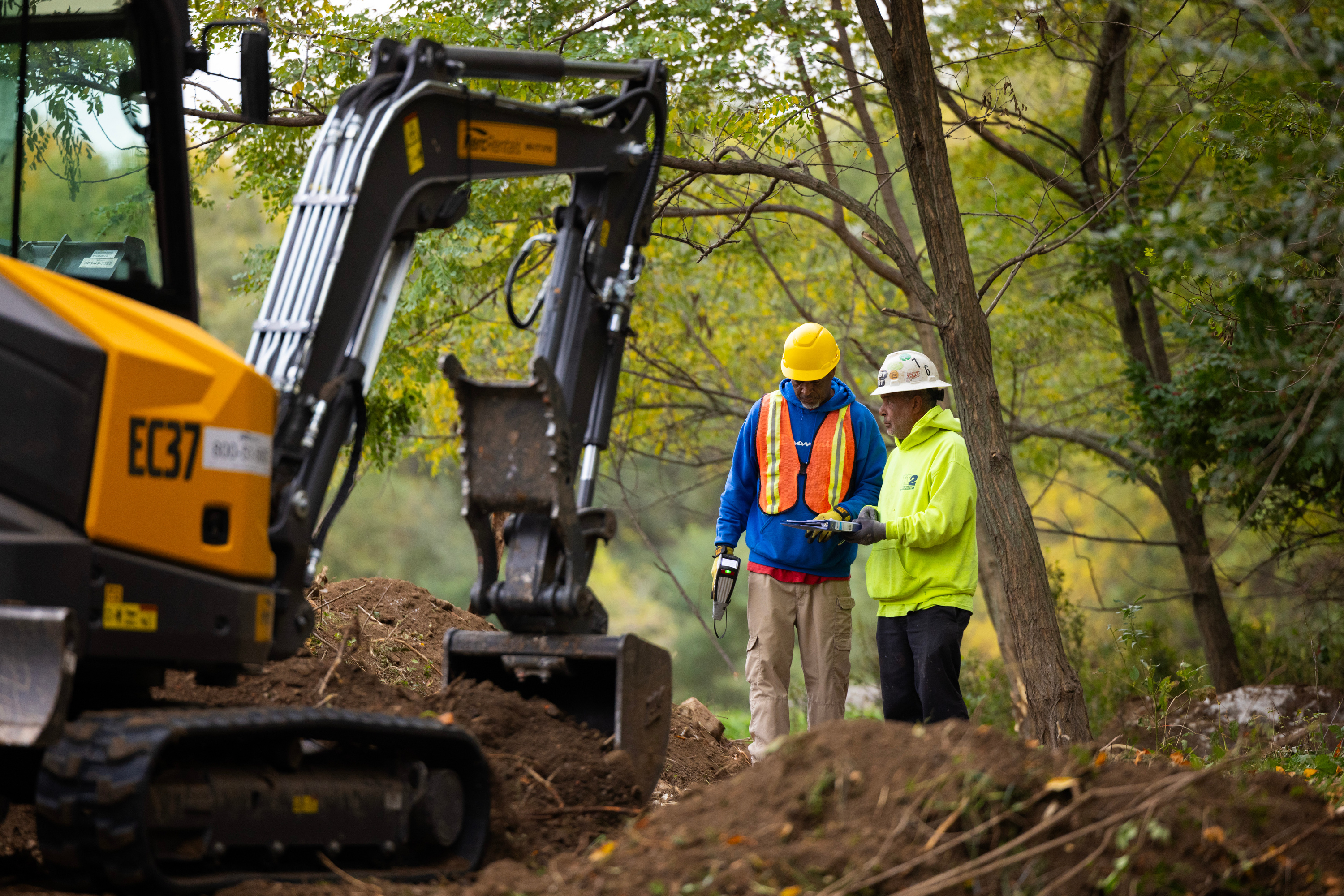 Two construction workers stand behind an excavator, surrounded by soil and trees.