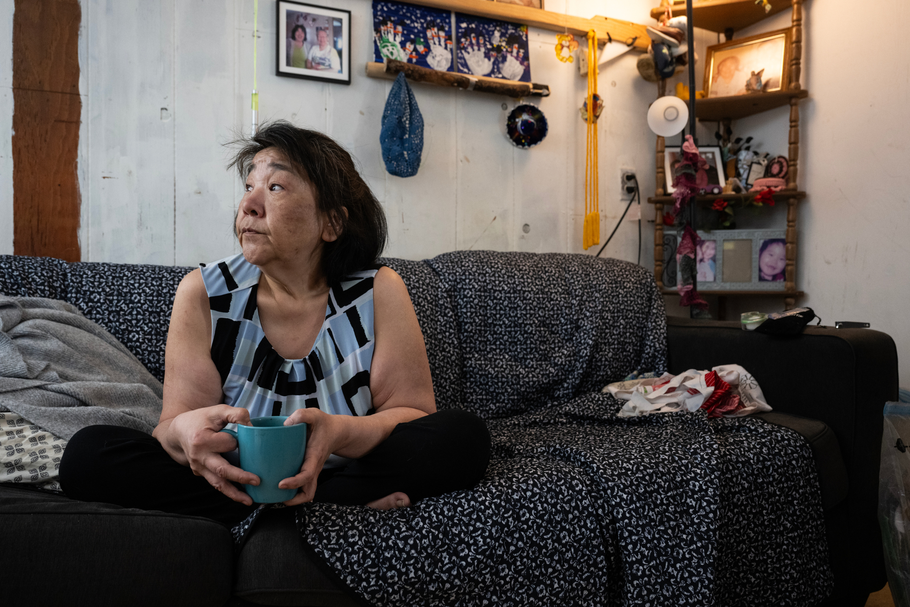 A woman looks off camera, holding a mug and sitting cross-legged on a couch covered in blankets. Behind her are framed family photos and knickknacks.