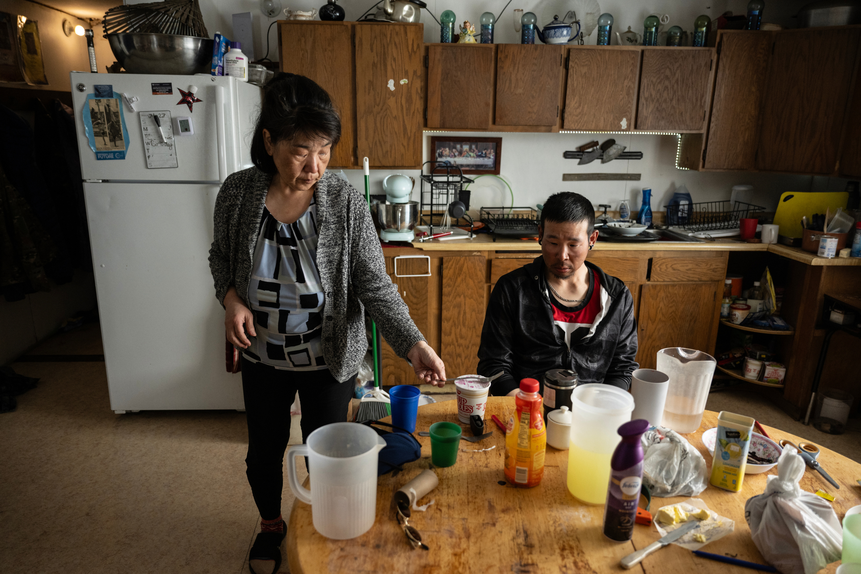 A standing woman places a fork on top of a styrofoam Cup Noodles container. A man sits at the kitchen table staring blankly at the dishes, cleaning supplies and food scattered across it. Behind the people is a row of wooden cabinets
