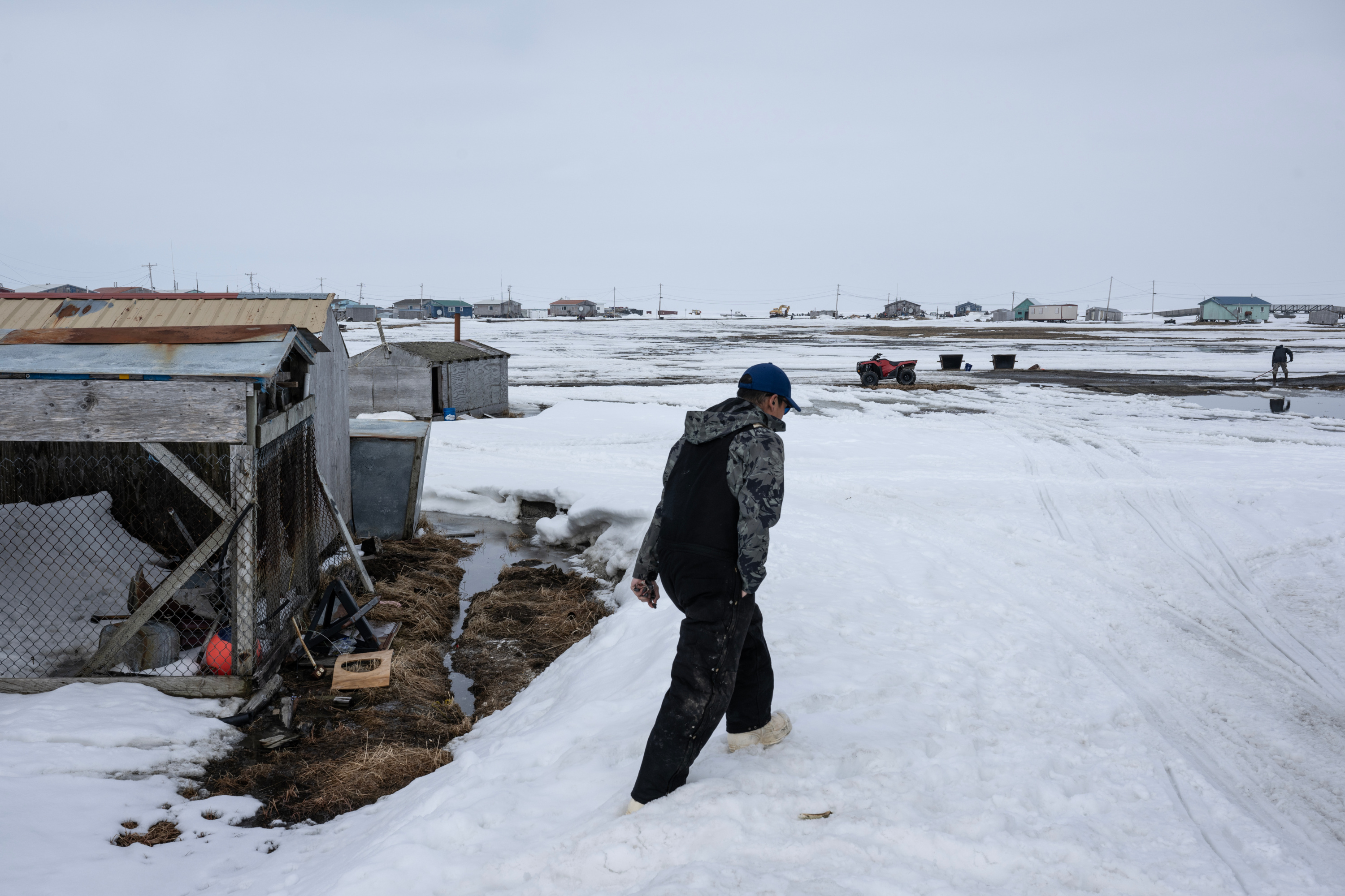 A man wearing black overalls, a hooded jacket and a baseball cap trudges through snow near small buildings and sheds.