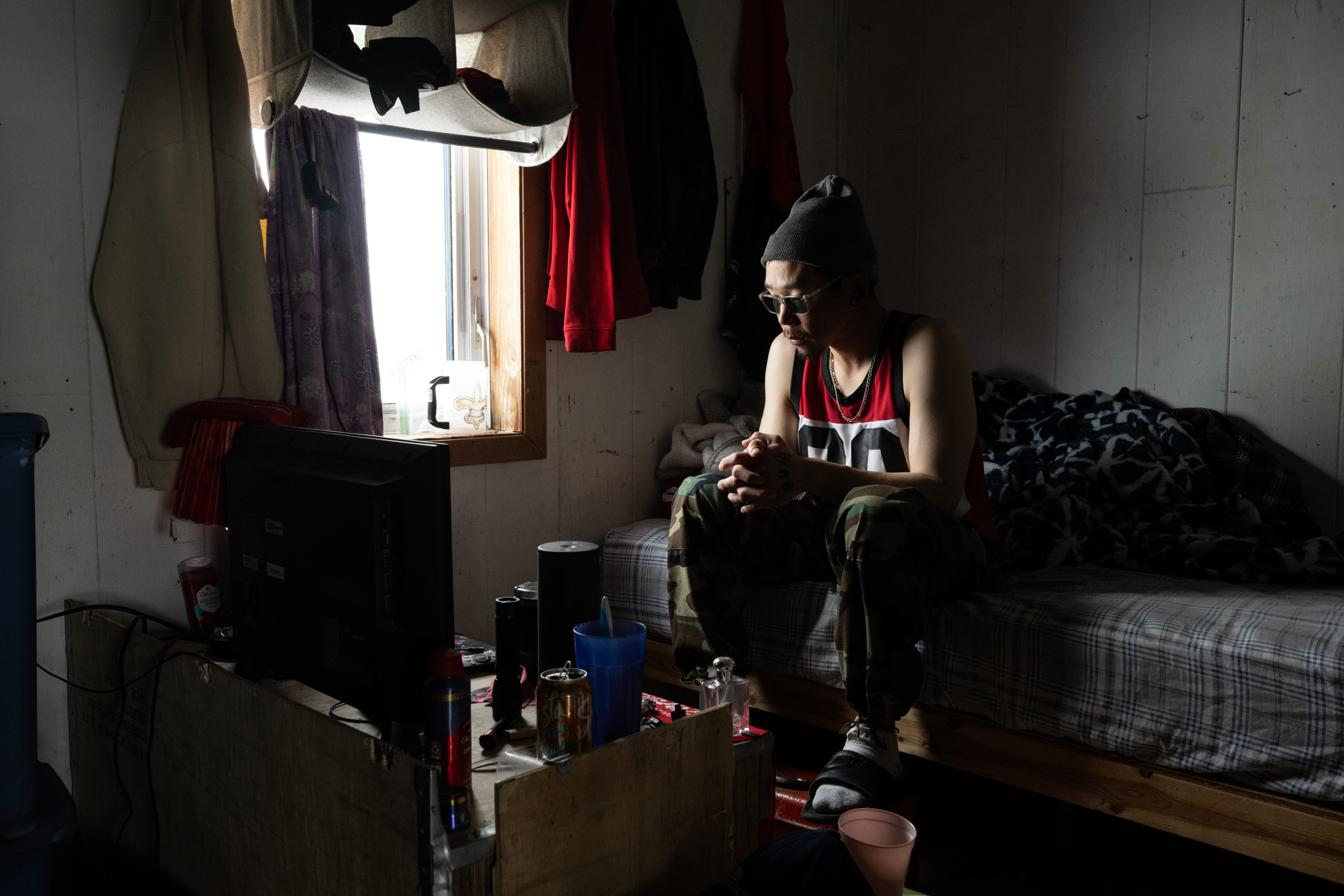 A man wearing a sleeveless sports jersey, a beanie and sunglasses sits on an unmade bed facing a television in a dim room lit only by a nearby window.