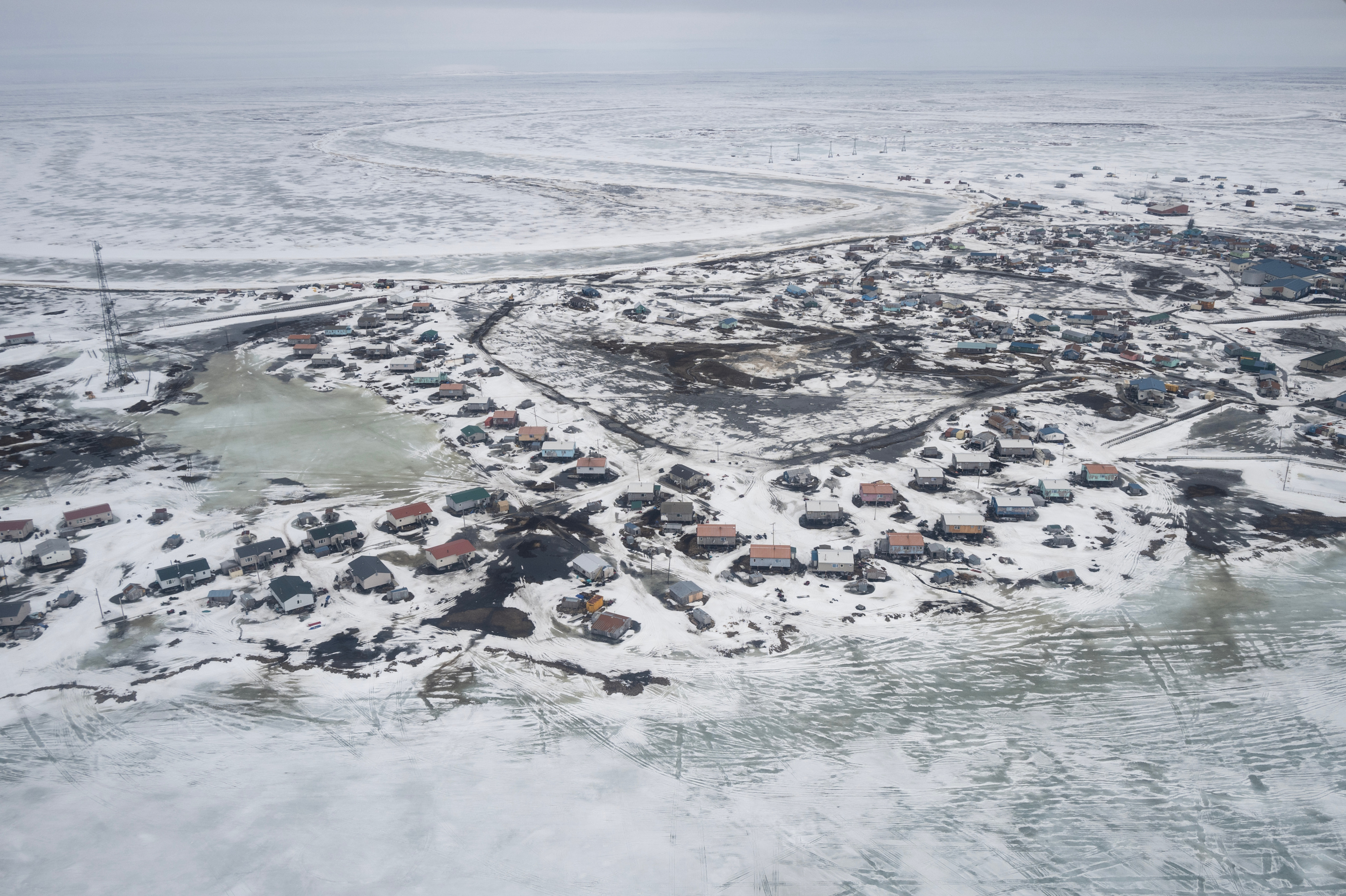 An aerial view of a snow-covered landscape. The foreground is dotted with houses, while a vast empty landscape stretches out in the background.