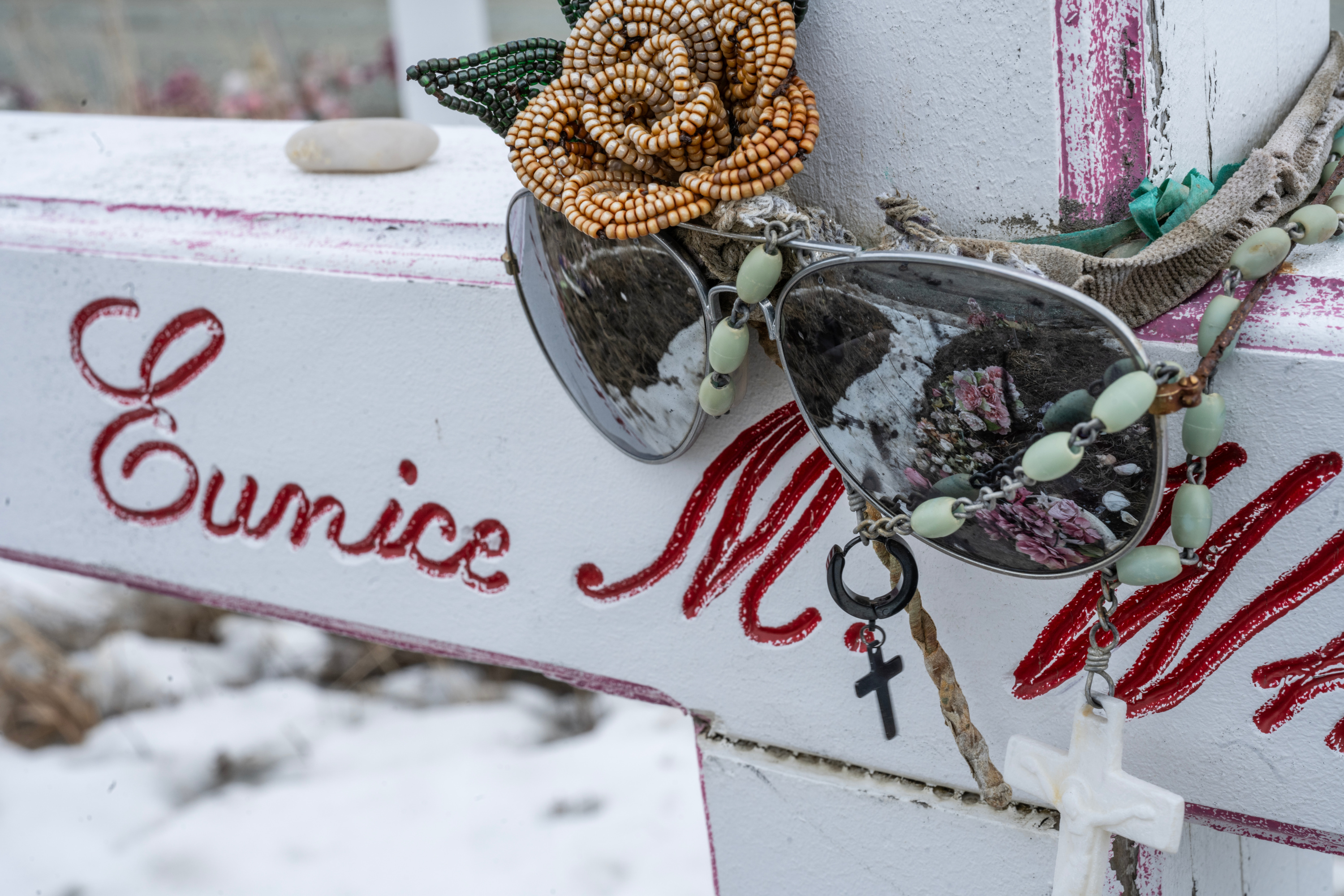 Close-up of a pair of aviator sunglasses, and a beaded necklace with a cross, and a beaded flower decorating a white cross emblazoned with the words “Eunice M. Whitman.”