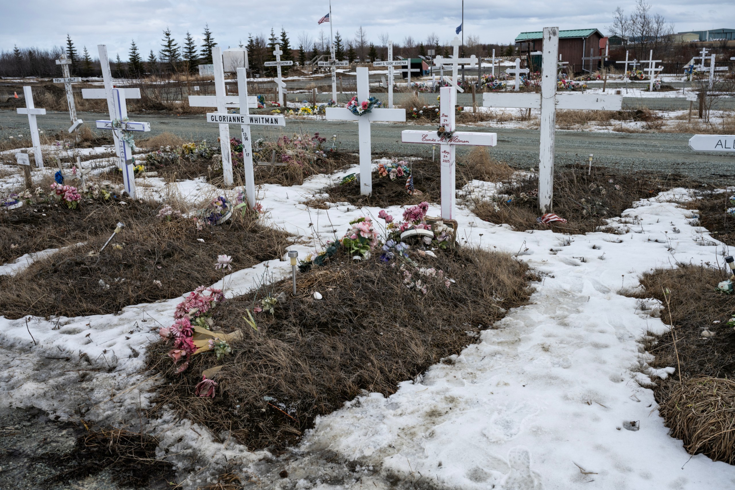 A row of graves with white crosses, piles of dirt and snow in the pathways. On the grave marked Eunice Whitman, bouquets of flowers lay on top of the soil.