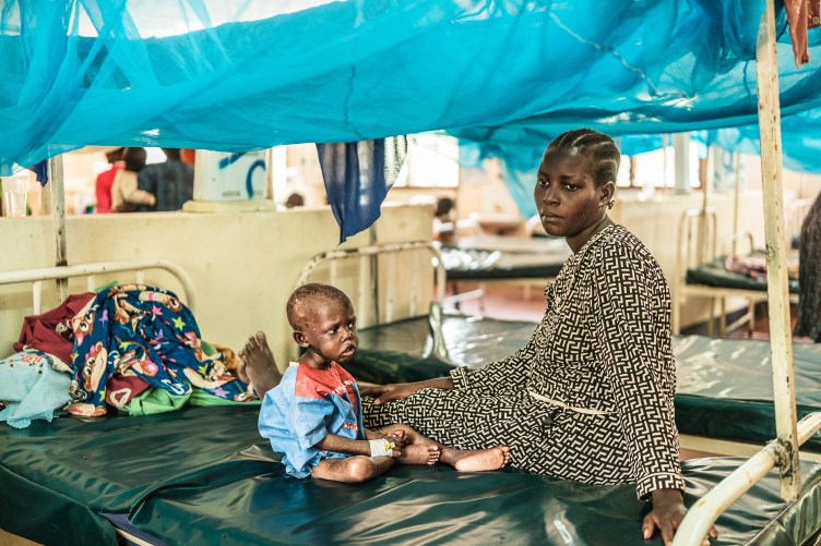 A woman in a patterned dress sits on a vinyl hospital bed with a small child in a too-big gown. Both have serious expressions. A mosquito net hangs over the bed.