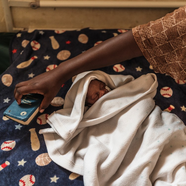 A photograph of an infant wrapped in blankets, lying on a bed with baseball-theme sheets. A woman’s hand reaches over the baby to pick up the Bible lying on the bed.